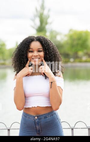 Smiling young black lady in summer clothes near lake and green trees in park looking at camera while touching face with finger in sunny day Stock Photo