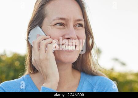 Happy adult Woman in casual outfit talking on smartphone while standing near green plants in park in sunny day Stock Photo