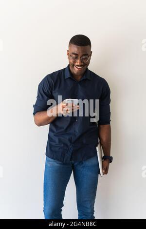 African American adult male in stylish outfit and eyeglasses standing with netbook in hands while using cellphone on white background Stock Photo