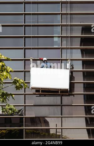 Back view of unrecognizable man in helmet and uniform in construction on ropes while washing windows of building on city street in daytime Stock Photo