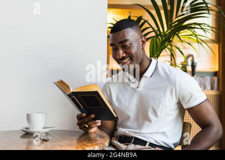Happy black adult male in casual clothes sitting at table in light cafeteria while drinking coffee and reading novel near cup of drink Stock Photo