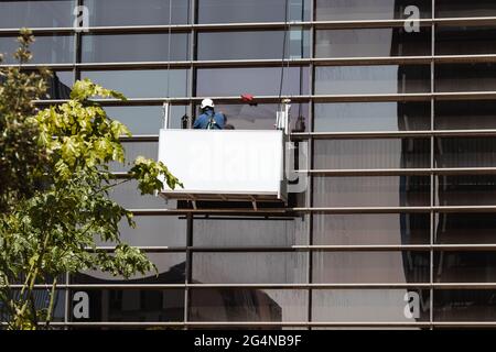 Back view of unrecognizable man in helmet and uniform in construction on ropes while washing windows of building on city street in daytime Stock Photo