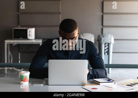 African American adult male in formal clothes and eyeglasses sitting at table and browsing on tablet near netbook and documents with takeaway drink in Stock Photo