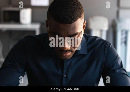 African American adult male in formal clothes and eyeglasses sitting at table and browsing on tablet near netbook and documents with takeaway drink in Stock Photo