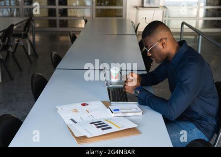 Side view of African American concentrated male in eyeglasses and formal clothes sitting in chair at table while using phone and netbook near document Stock Photo