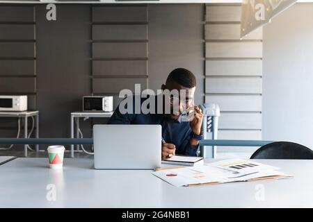 African American male in eyeglasses and formal clothes at table having conversation on cellphone and taking notes in notepad near netbook and document Stock Photo