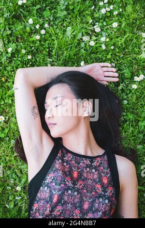 Overhead view of gentle ethnic female in ornamental wear lying with closed eyes on lawn with blooming chamomiles in summer Stock Photo