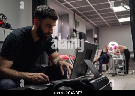 Thoughtful adult guy in casual outfit with tattoos sketching on computer in bright tattoo studio near tattooist making tattoo for client Stock Photo