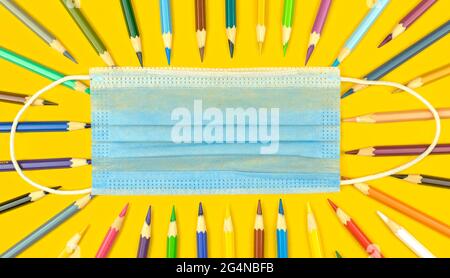 Back to school during coronavirus pandemic, concept of education with medical mask, color pencils on the yellow desktop, top view Stock Photo