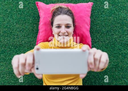 From above of positive adult female in casual clothes lying on pillow on green carpet while watching interesting video on mobile phone Stock Photo