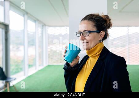 Positive adult female in stylish outfit with mug with coffee in light room looking away near windows and column Stock Photo