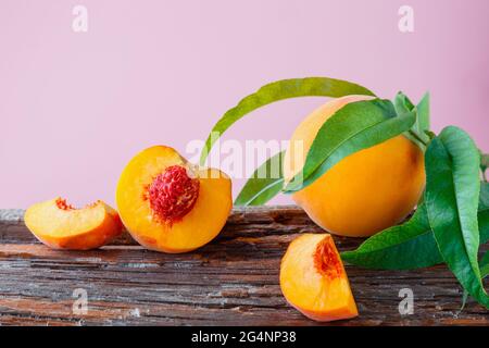 Peaches with leaves on wooden board. Peach in halves with bone, chopped pieces wedges peach halves. Fresh organic peaches fruit on color pink Stock Photo