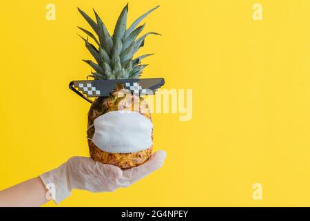 Funny pineapple face in a protective medical mask. Doctor hand in rubber glove holds summer tropical fruit pineapple in sunglasses. Safe travel Stock Photo