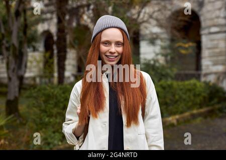 Close-up Portrait Of Relaxed Woman With Natural Red Hair On Travel Trip, Outdoors, Posing At Camera, Smiling, In Coat And Hat. Old Building In The Bac Stock Photo