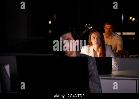 redhead female programmer working on new project, sit in shock by seeing the time. Hardworking woman working late at night at office with colleagues. Stock Photo