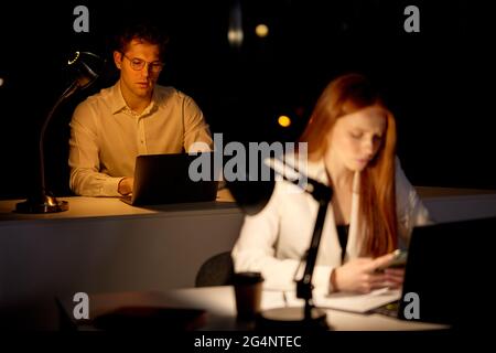 Tired Young Capable Manager Working Late Hours in Office. Office is Dark. Male is working on computer with colleagues. business, deadline, work concep Stock Photo
