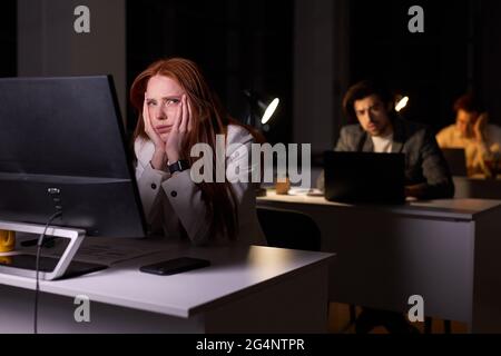 Sleepy caucasian female looking at side while sitting at desk in office at night, woman wants to go home, leaned on hands, dressed in formal wear. wor Stock Photo