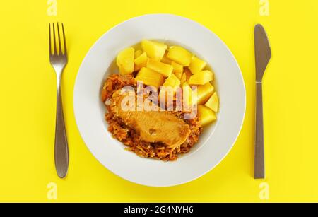 Baked potatoes, pork meat and cabbage in white plate on yellow board with cutlery, view form above Stock Photo