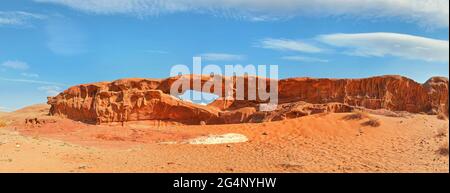 Little arc or small rock window formation in Wadi Rum desert, bright sun shines on red dust and rocks, blue sky above, high resolution wide panorama Stock Photo