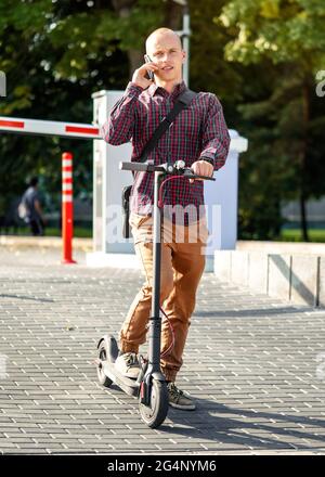 Young man in casual trousers and shirt standing next to his electric scooter, talking on the mobile phone Stock Photo