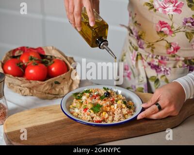 woman's hands drizzling olive oil on plate of quinoa salad in home kitchen, paper bag bottom with tomatoes and white kitchen drawers Stock Photo