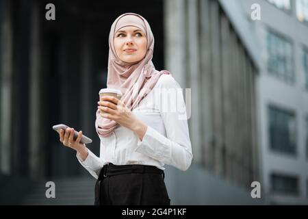 Attractive arabian woman standing near office center and holding modern smartphone and cup of coffee. Confident lady in formal clothes and hijab looking aside while posing on street. Stock Photo