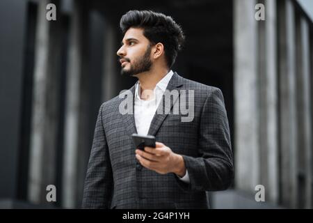 Successful arabian businessman standing on city street, holding modern cell phone and looking aside. Handsome man with beard wearing stylish formal clothes. Stock Photo