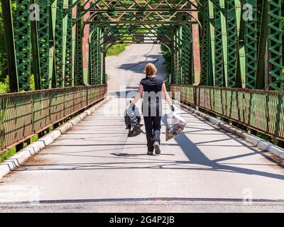 Middle aged Caucasian woman with red hair, black clothes and gloves, carries plastic and a garbage bag across a metal bridge for a volunteer clean up. Stock Photo