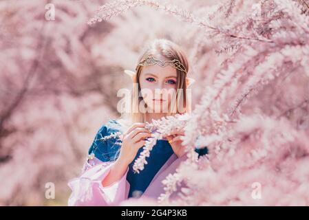 Beautiful romantic blonde girl with blue eyes posing in forest. Dreaming fairytale girl. Cosplay character. Portrait of elf girl with long ears touche Stock Photo