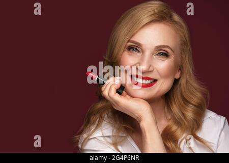 Portrait of a beautiful elderly woman in a white shirt with classic makeup and blond hair with red lipstick in hand Stock Photo