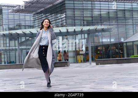 Young beautiful girl walking outside office building using mobile phone and happily calling customers