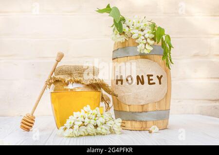 Fresh liquid acacia honey harvest 2021 in a wooden barrel and a transparent jar with a canvas napkin on the lid. Branches of flowering white acacia, r Stock Photo