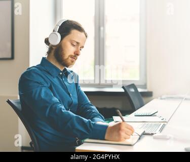 Male office worker dressed in casual clothes in headphones watching online webinar on laptop Stock Photo