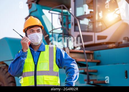Portrait of warehouse asian man worker driver truck driver crane lifting up container with safety equipment standing in front of big forklift use radi Stock Photo