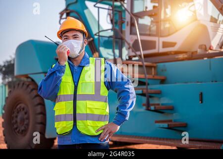 Warehouse asian man worker driver truck driver crane lifting up container with safety equipment, helmet is standing in front of big forklift use radio Stock Photo