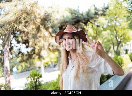 Cheerful blond woman in casual style wearing clothes and felt hat looks into the camera and shows the sign Peas outdoor Stock Photo