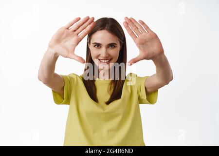 Just imagine. Smiling creative young woman look through hand frames with determined grin, found perfect angle, making shot, standing against white Stock Photo