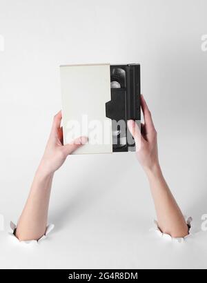 Female hands hold a video cassette in a case through the torn holes of a white background. Creative art. Retro style Stock Photo