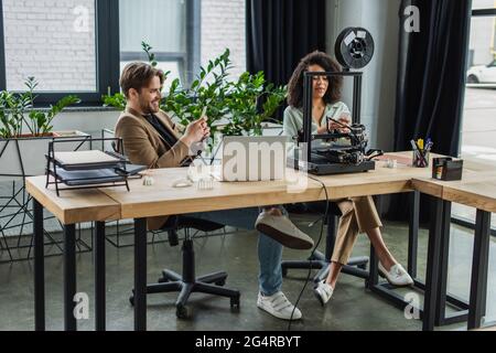 smiling interracial colleagues messaging on cellphones near 3D printer and laptop in modern office Stock Photo