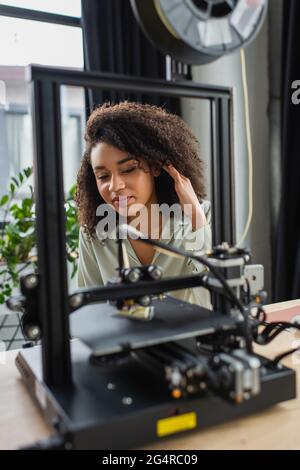 smiling young african american woman looking at 3D printer and adjusting hair in modern office Stock Photo