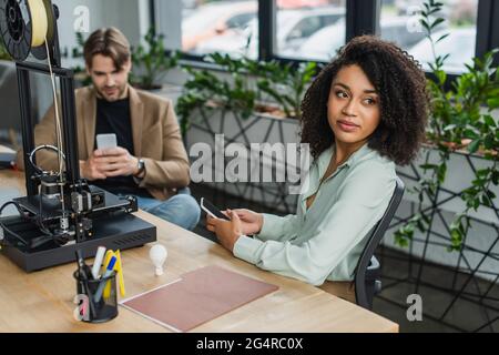 interracial colleagues sitting with cellphones in hands near 3D printer in modern office Stock Photo