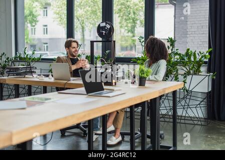 smiling interracial colleagues sitting with cellphones in hands near 3D printer and laptops in modern office Stock Photo