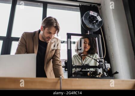 concentrated young interracial designers working with 3D printer near laptop in modern office Stock Photo