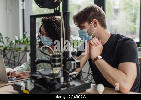 interracial colleagues in protective masks working with laptop near 3D printer in modern office Stock Photo