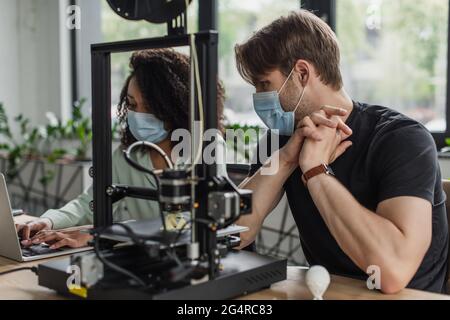 interracial colleagues in medical masks working with 3D printer and laptop in modern office Stock Photo