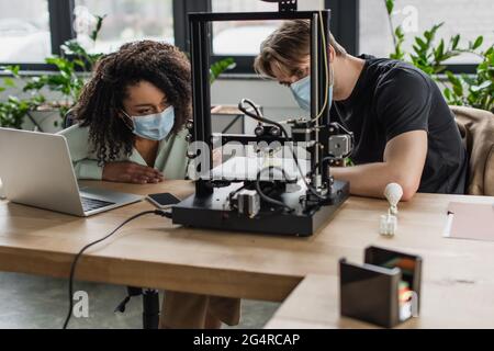 interracial colleagues in medical masks looking at 3D printed model production in modern office Stock Photo