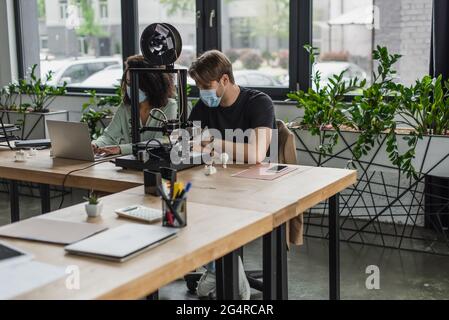 interracial colleagues in medical masks typing using laptop near 3D printer in modern office Stock Photo