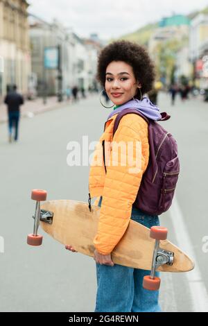 Smiling african american woman with backpack and longboard standing on urban street Stock Photo