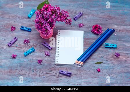 Blank notebook, pencils, lilac flowers and colorful pegs on vintage wooden painted blue background. Stock Photo