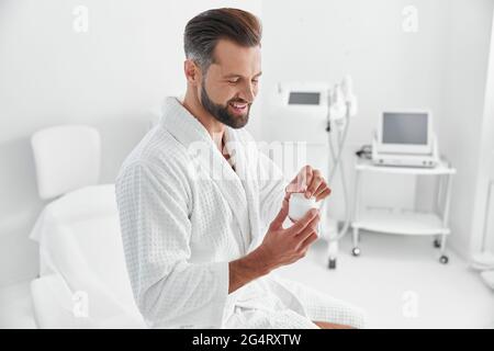 Grey haired person takes cream from open jar sitting on couch in cosmetologycal clinic Stock Photo
